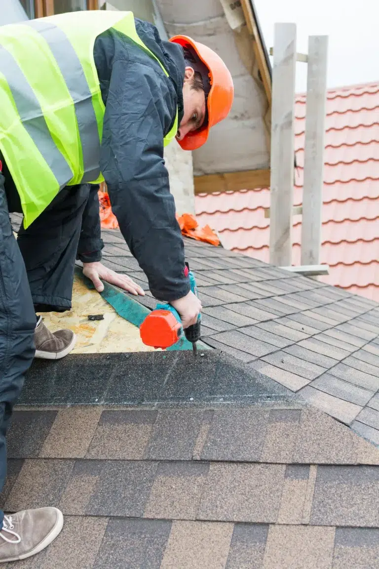 A roofer working in uniform using air or pneumatic nail gun and installing shingles on top of a slanted roof